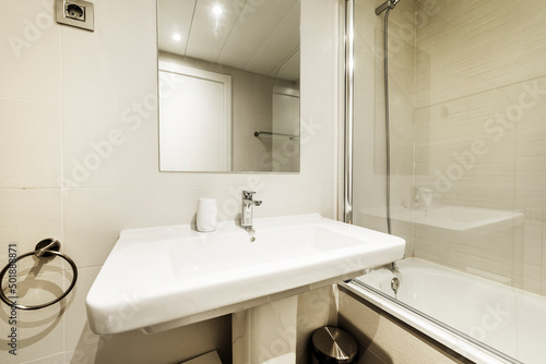 Bathroom with white porcelain sink on porcelain pedestal, frameless mirror, glass-enclosed tub, and light tile