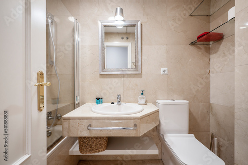 Bathroom with cream marble countertop to match the walls  mirror with silver frame and shower cabin with glass partition