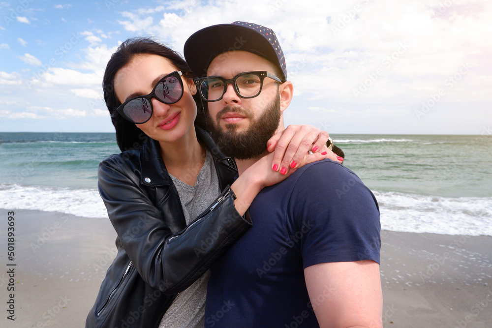 Beautiful young couple of hipsters in fashionable clothes on the background of the sea.Bearded guy, t-shirt, blue baseball cap, wearing sunglasses, smiling, happy lovers, on the beach by the sea