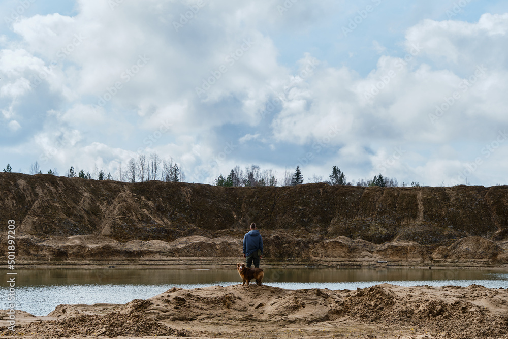 Sand quarry with water. Caucasian man travels with Australian Shepherd dog. Person stands on the sandy shore of river or lake and looks into distance view from behind.