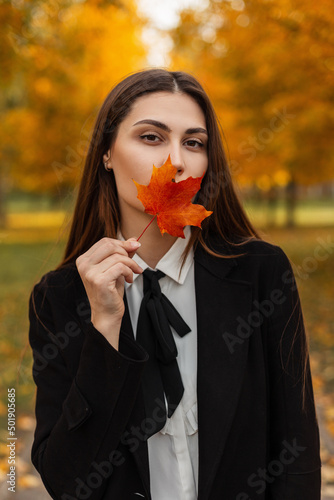 Fashion beautiful young brunette woman in stylish business clothes walks in colored autumn park and covers face with a red-orange fall maple leaf