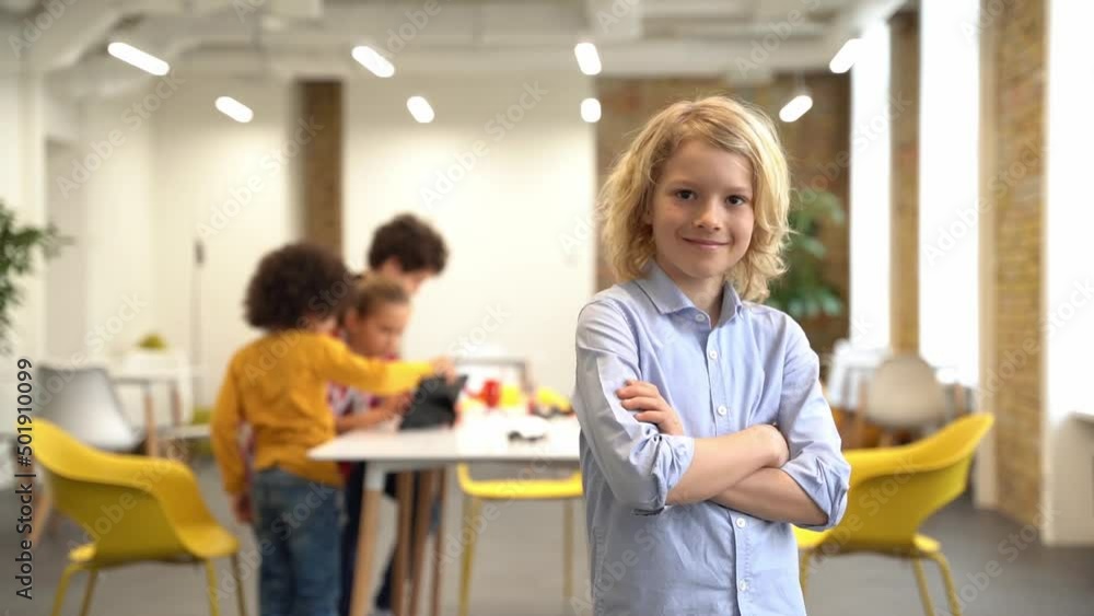 Smart guy. Joyful boy smiling at camera, standing with arms crossed while posing for camera during crafting classes. Kids working on diy robot in the background Smart guy. Joyful boy smiling at camera, standing with arms crossed while posing for camera during crafting classes. Kids working on diy robot in the background