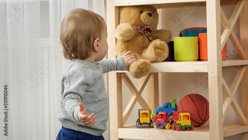 Cute baby boy taking teddy bear toy from bookshelf in playroom. Child education photo