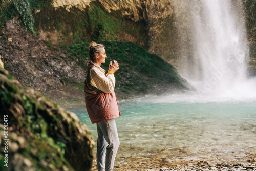 A young woman drinking a hot drink in a mountain canyon in a nature reserve near a large waterfall.