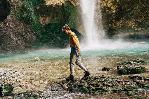 A young active female tourist at a large mountain waterfall steps through a stream.