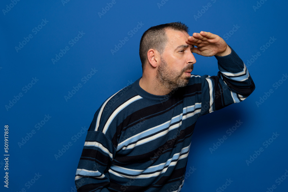Cheerful bearded young man in striped pullover posing isolated on blue background in studio. People lifestyle concept. Mock up copy space. Holding hand to forehead looking into the distance.