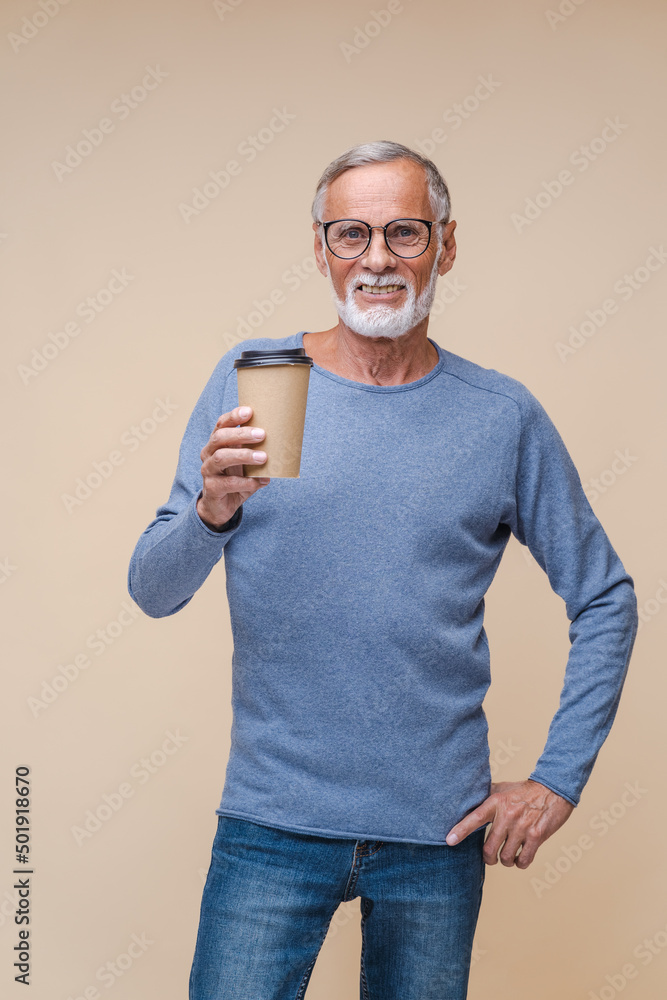 Joyful elderly man in black-rimmed glasses holds paper cup of coffee smiling. Stylish bearded pensioner enjoys hot drink standing on beige background Joyful elderly man in black-rimmed glasses holds paper cup of coffee smiling. Stylish bearded pensioner enjoys hot drink standing on beige background