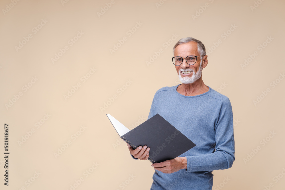 Happy senior man holds big notepad with hands smiling widely. Positive bearded businessman in black-rimmed spectacles stands on beige background