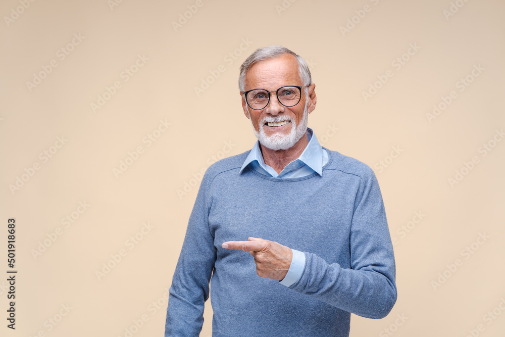 Happy bearded elderly man businessman in glasses makes hand gesture pointing index finger. Senior person in sweater smiles standing on beige background Happy bearded elderly man businessman in glasses makes hand gesture pointing index finger. Senior person in sweater smiles standing on beige background