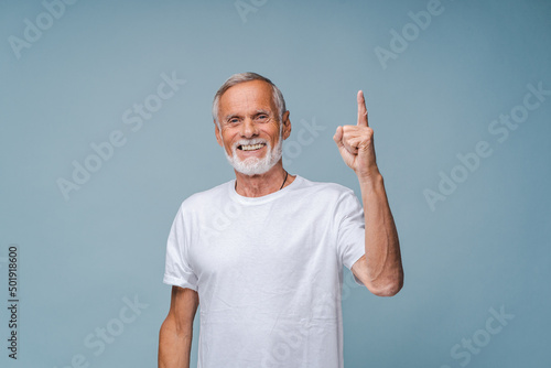 Positive elderly man in white t-shirt points up index finger smiling. Grey-haired senior male person shows finger gesture standing on blue background