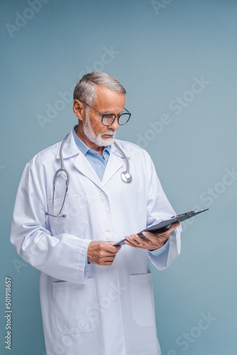 Experienced senior doctor in glasses holds clipboard checking schedule. Focused bearded man with stethoscope in coat reads patient medical history at blue wall
