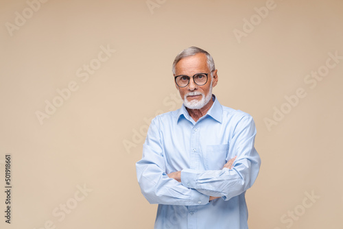 Serious elderly man in black-rimmed glasses stands with arms crossed looking in camera. Bearded male person in blue shirt on beige background