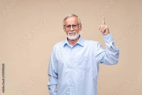 Cheerful elderly man in glasses points index finger up smiling. Bearded senior in light blue shirt grandfather shows gesture standing on beige background © Alla