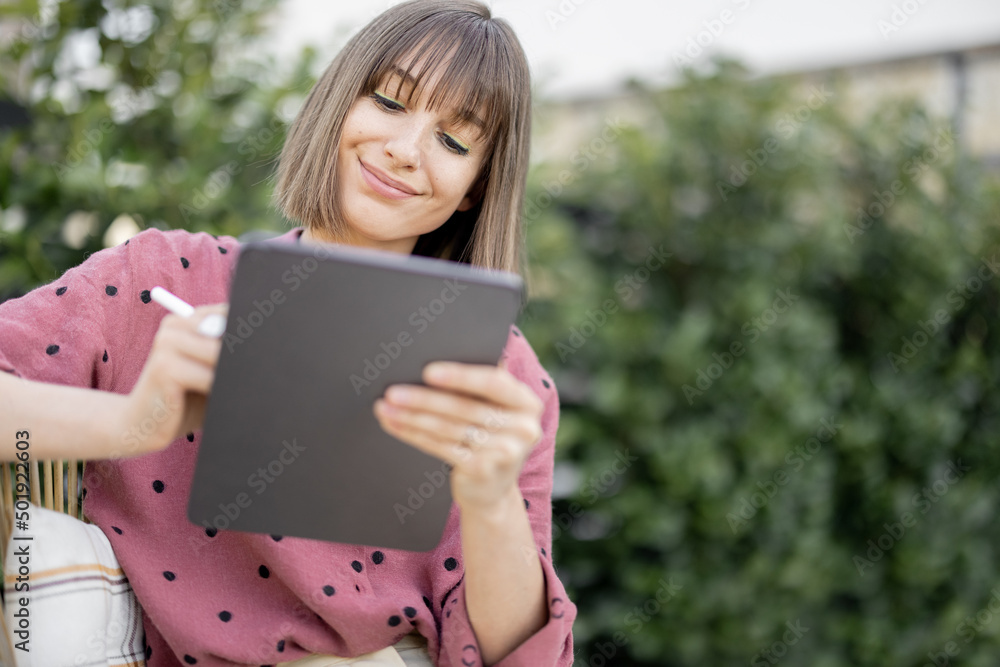 Portrait of young creative woman works with stylus on a digital tablet at backyard outdoors. Concept of remote work or leisure time with gadgets at cozy atmosphere Portrait of young creative woman works with stylus on a digital tablet at backyard outdoors. Concept of remote work or leisure time with gadgets at cozy atmosphere