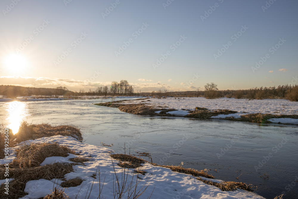 March sunny evening by the river. Blue sky over the horizon. A picturesque landscape, early spring, a river with snow-covered banks, dry grass and bushes. The first thaws, the snow is melting March sunny evening by the river. Blue sky over the horizon. A picturesque landscape, early spring, a river with snow-covered banks, dry grass and bushes. The first thaws, the snow is melting