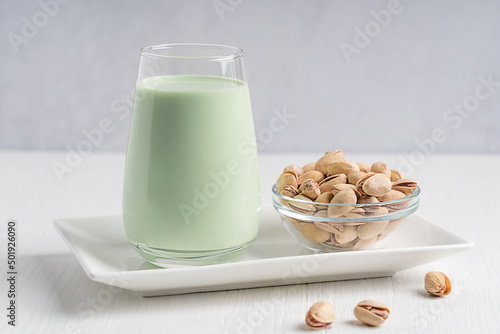 Natural plant based pistachio milk with creamy flavor and green color, lactose free, suitable for vegetarian or vegan diet served in glass with bowl of seeds on white wooden table for breakfast