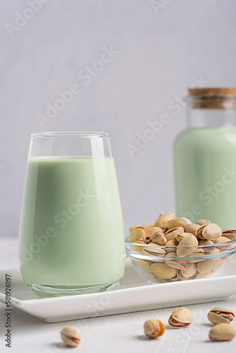 Healthy organic non-dairy pistachio milk with creamy flavor and green color, lactose free, suitable for vegetarian or vegan diet served in drinking glass with bowl of seeds on white wooden table