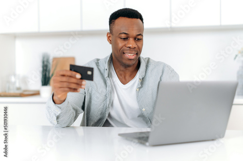 Happy african american man sitting at home, dressed in casual wear, using laptop and credit card for online shopping, entering data for payment, online transaction, order home delivery, smiling