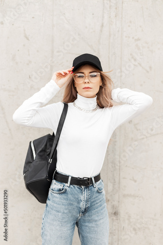 Fashion young woman with glasses in casual trendy clothes with black cap, white sweatshirt, jeans and bag stands near a gray concrete wall