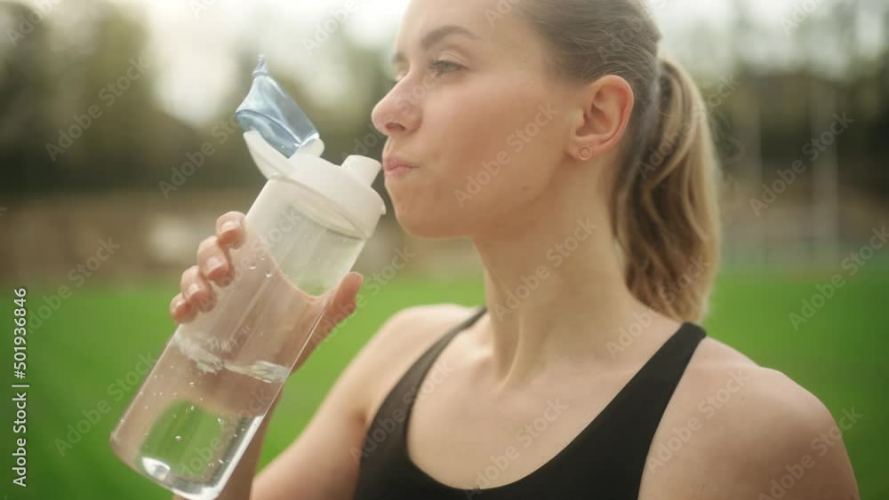Attractive blond young woman drinking from sport bottle during a workout pause in stadium. Fitness woman drinking water from bottle after working out. . High quality 4k footage Attractive blond young woman drinking from sport bottle during a workout pause in stadium. Fitness woman drinking water from bottle after working out. . High quality 4k footage