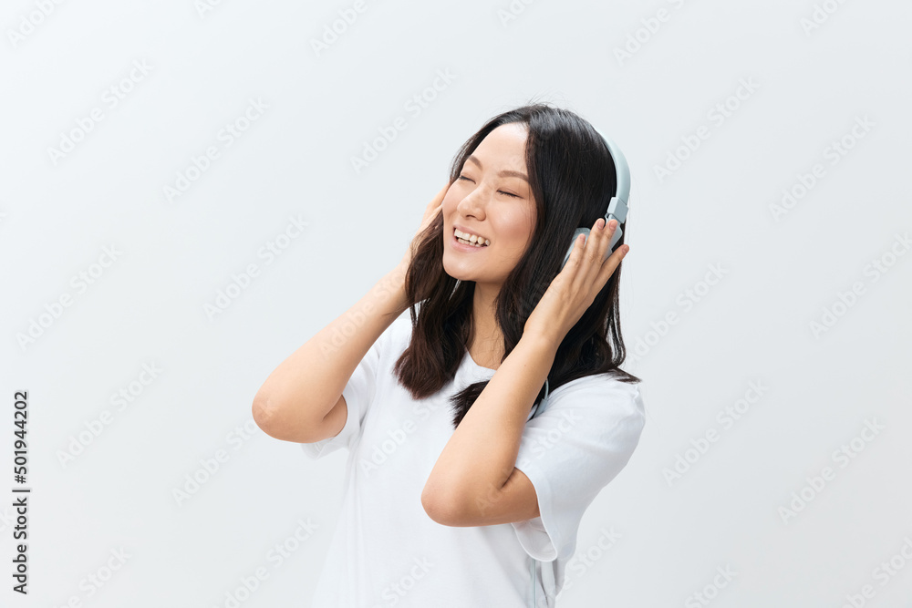 Relaxing happy cute Asian young female in white basic t-shirt touch headphones closing eyes posing isolated on over white studio background. The best offer for ad. Favorite Music App Ad concept Relaxing happy cute Asian young female in white basic t-shirt touch headphones closing eyes posing isolated on over white studio background. The best offer for ad. Favorite Music App Ad concept
