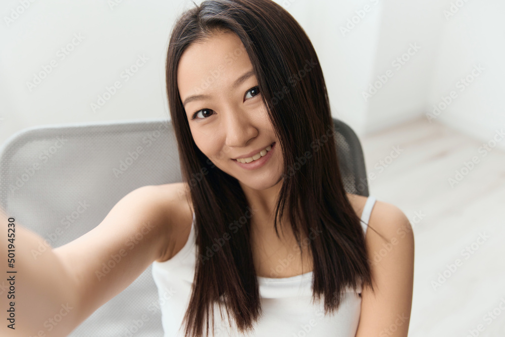 Smiling happy tanned lovely young Asian woman doing selfie sit on chair at home interior living room. Distance communication, Remote work, Social media concept. Cool offer Wide angle Selfie view Smiling happy tanned lovely young Asian woman doing selfie sit on chair at home interior living room. Distance communication, Remote work, Social media concept. Cool offer Wide angle Selfie view