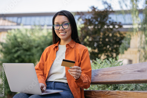 Smiling confident asian business woman holding credit card using laptop computer shopping online outdoors. Young stylish freelancer receive payment sitting on bench. Successful business