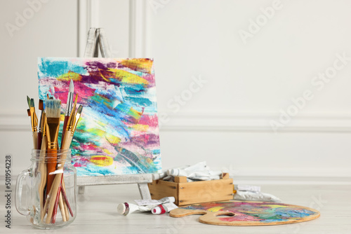 Different brushes, easel with abstract picture and paints on white wooden table. Space for text