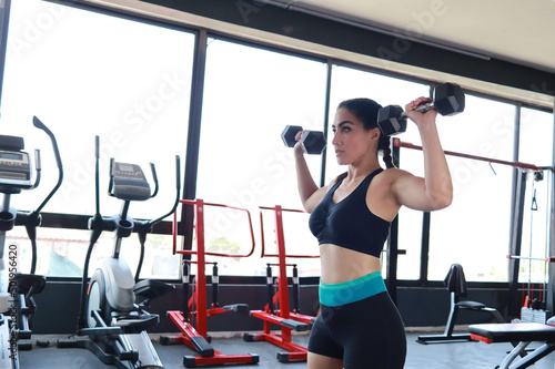 Beautiful woman exercises her arms lifting dumbbells in the gym alone determined sporty healthy in daylight with black sportswear.