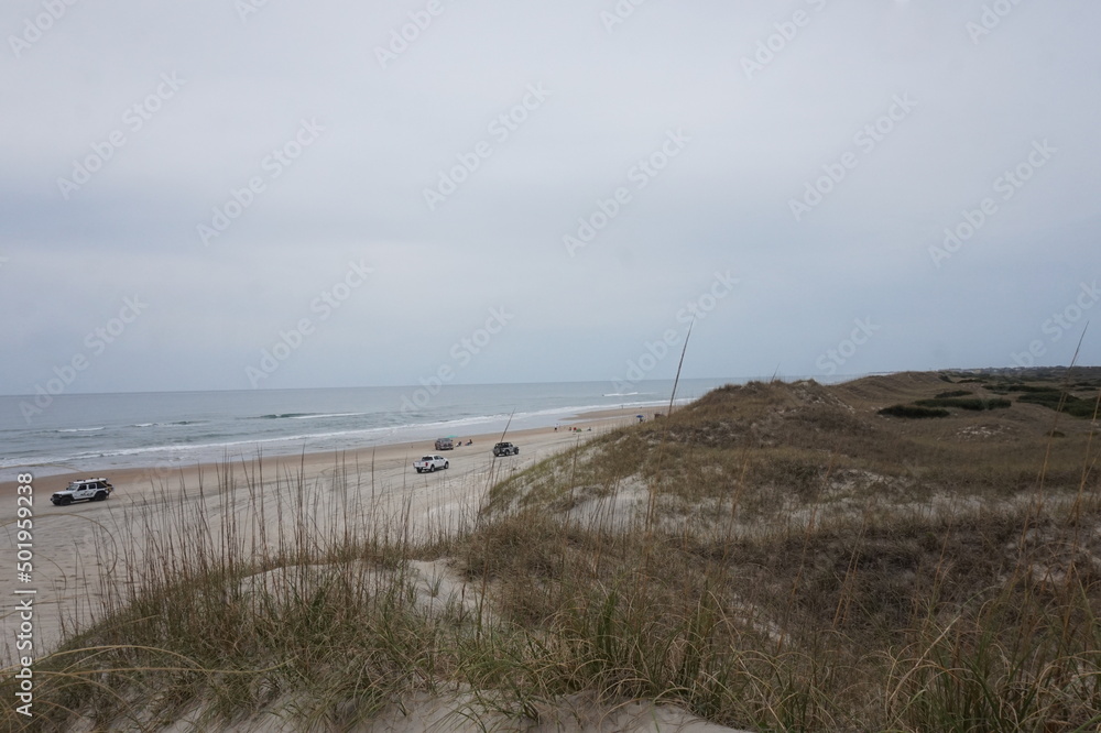 Landscape of Cars, Pickups on Beach with Ocean Water in Daylight fron Top of Grassy Dunes
