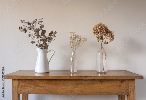 Close up of dried flowers and leaves in glass bottles and white jug on oak side table against beige wall (selective focus)