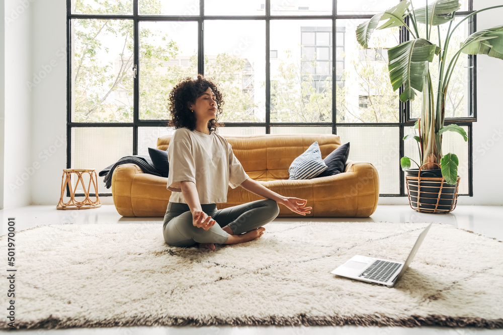 Young multiracial latina woman meditating at home with online video meditation lesson using laptop. - obrazy, fototapety, plakaty 