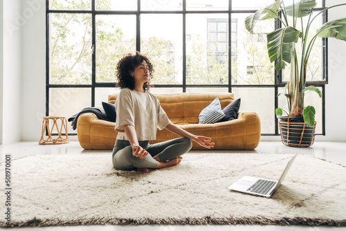 Young multiracial latina woman meditating at home with online video meditation lesson using laptop.