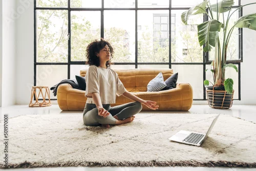 Fototapeta na wymiar Young multiracial latina woman meditating at home with online video meditation lesson using laptop.