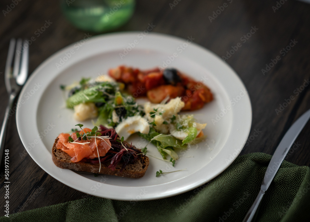 One plate dish on a dark brown surface with a moss green cloth. Boiled chicken tomato, Nicoise-style salad and smoked salmon open sandwich.