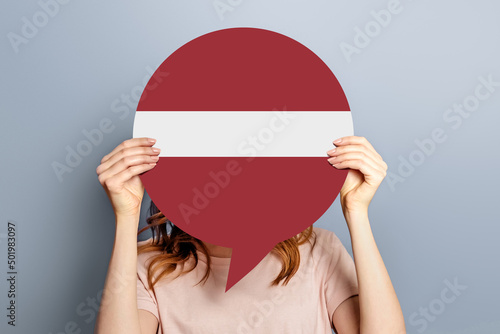 Voting in latvia concept. Student girl holds blank white speech bubble with latvia flag isolated over grey background. woman holding a round information piece of paper dialog
