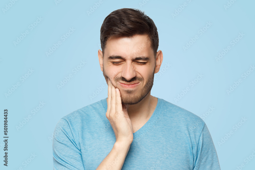 Young man with closed eyes suffering from severe toothache, touching jaw with fingers