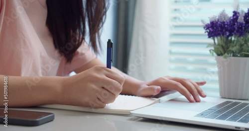 An woman Asian university college student uses a laptop computer on the table at his home.Female hands using a laptop and take notes on notepad.for studying online.Online education concept.