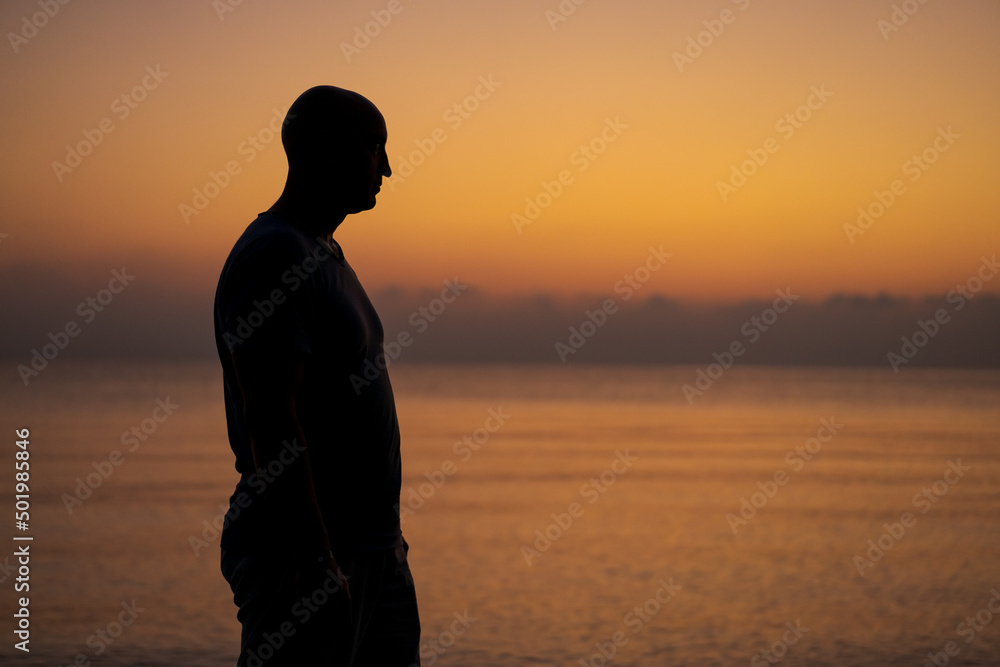 Male silhouette on background sea at sunset looks to side alone. Man enjoying a warm evening on the shore in the evening when it gets dark, reflects and dreams