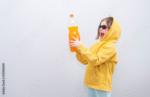 Child, Caucasian 6 years old, in a yellow sweatshirt with a hood on a gray background. The child rejoices and gesture holds a bottle of lemonade in his hands.