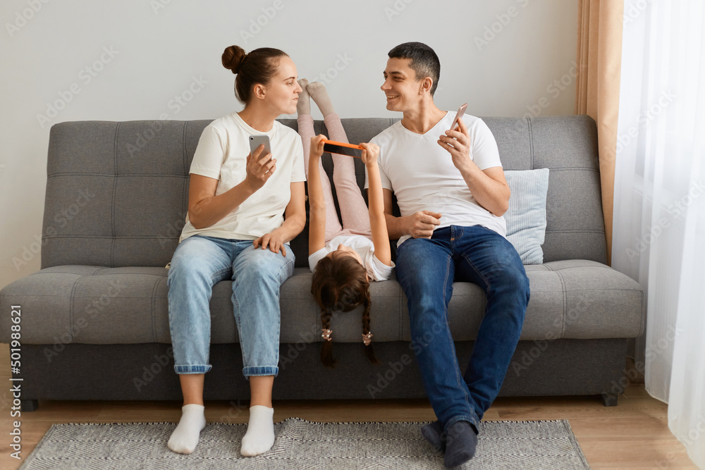 Portrait of young adult family of holding and using mobile phones while sitting on sofa. Parents and their daughters with modern gadgets, husband showing his device to her wife and smiling. Portrait of young adult family of holding and using mobile phones while sitting on sofa. Parents and their daughters with modern gadgets, husband showing his device to her wife and smiling.