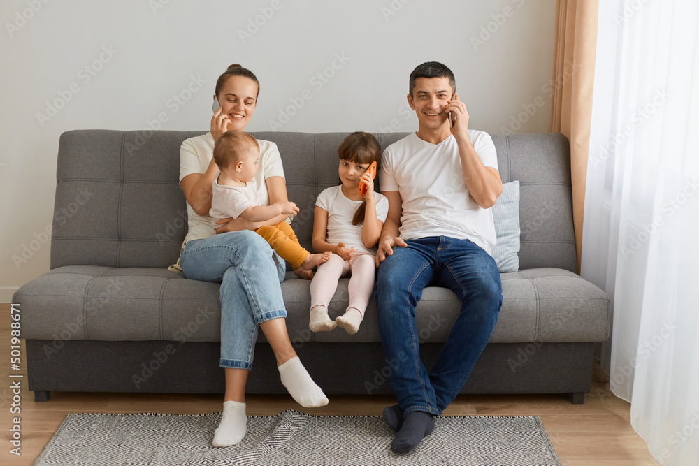 Horizontal shot of busy family sitting on sofa and having conversation on smart phone, looking at camera, kid with smart phone talking too, family spending time with gadgets at home. Horizontal shot of busy family sitting on sofa and having conversation on smart phone, looking at camera, kid with smart phone talking too, family spending time with gadgets at home.