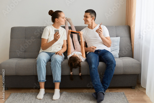 Portrait of young adult family of holding and using mobile phones while sitting on sofa. Parents and their daughters with modern gadgets, husband showing his device to her wife and smiling.