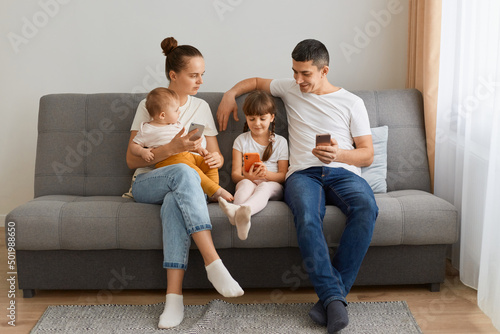 Portrait of attractive man and woman wearing casual attires sitting on coughwith cell phones and looking at her daughter's cell phone, contolling what kid watching in internet.