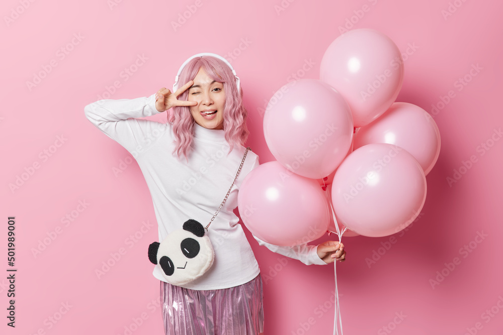 Good looking pink haired Asian girl makes peace gesture over eye sticks out tongue foolishes around on party dressed festively holds bunch of inflated helium balloons poses for making photo.