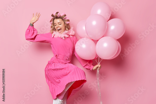 Happy cheerful young woman wears festive dress dances against pink background with bunch of balloons applies hair rollers for making hairstyle has positive expression. Special occasion concept