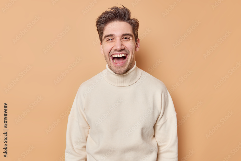 Waist up shot of overjoyed handsome man laughs happily expresses sincere emotions hears something funny dressed in casual white jumper isolated over brown background. People and happiness concept Waist up shot of overjoyed handsome man laughs happily expresses sincere emotions hears something funny dressed in casual white jumper isolated over brown background. People and happiness concept