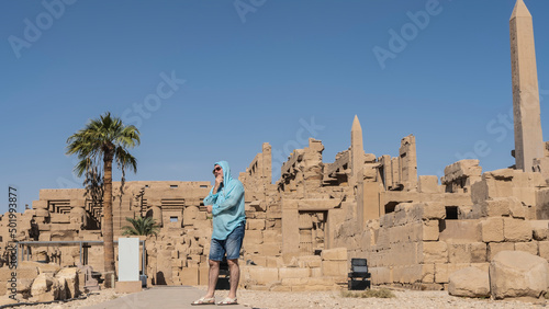 A man stands in the ruins of the ancient Karnak temple of Luxor, looking into the distance. The hand holds the chin. The face is thoughtful. Dilapidated stone walls, obelisks against a blue sky. Egypt