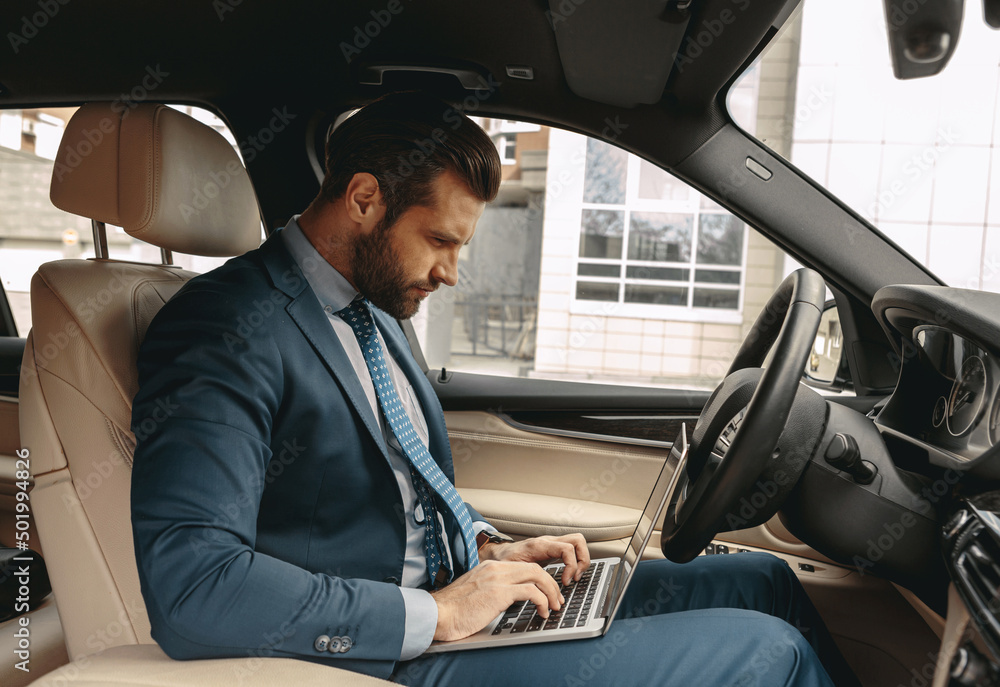 Elegant business man in suit typing something on laptop in the car