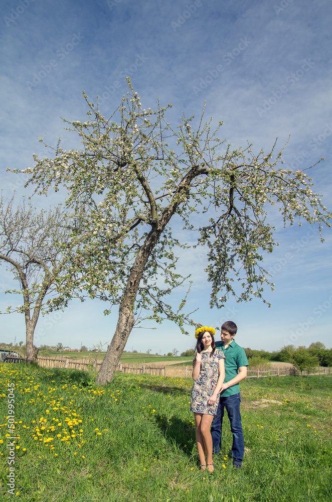 Loving couple near a blossoming tree on a background of spring blue sky. Girl with a wreath of dandelions on her head.